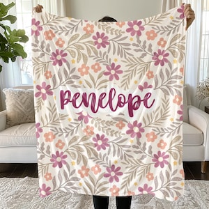 May include: A soft throw blanket featuring a repeating pattern of pink and peach flowers with grey leaves on a white background. The name "penelope" is printed in a dark pink cursive script across the center of the floral design.