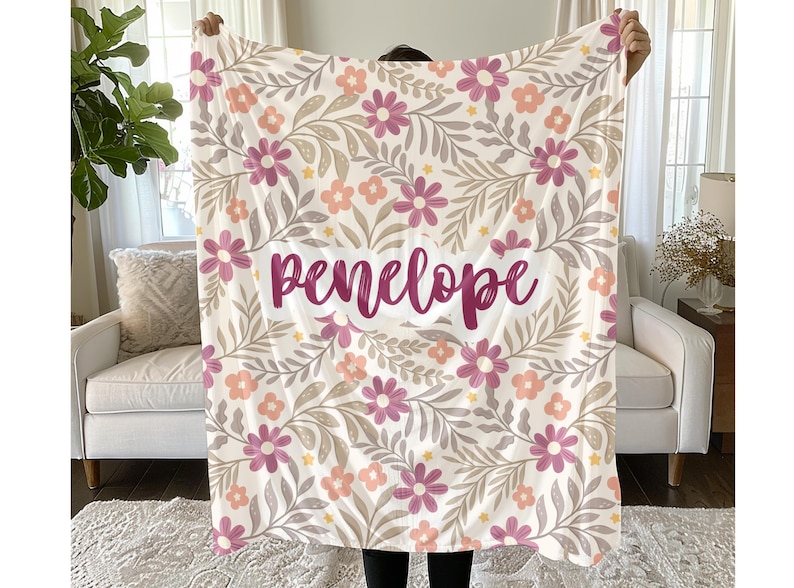 May include: A soft throw blanket featuring a repeating pattern of pink and peach flowers with grey leaves on a white background. The name "penelope" is printed in a dark pink cursive script across the center of the floral design.