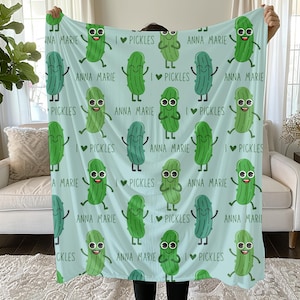 May include: A light blue blanket featuring a repeating pattern of cartoon pickles with smiling faces and the text "I Pickles" and "Anna Marie". The blanket is held up.