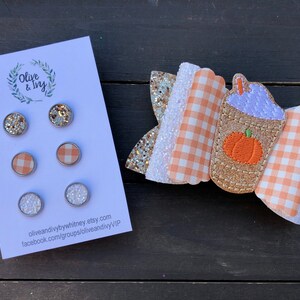 Op de afbeelding: Set van drie 2,5 cm ronde haarstrikken met een pompoenspeceratte-ontwerp. De strikken zijn gemaakt van oranje en wit gingham-stof, gouden glitterstof en een paarse en oranje pompoenspeceratte-applicatie. De set bevat ook drie paar oorbellen met gouden glitter, witte glitter en oranje en wit gingham-stof.