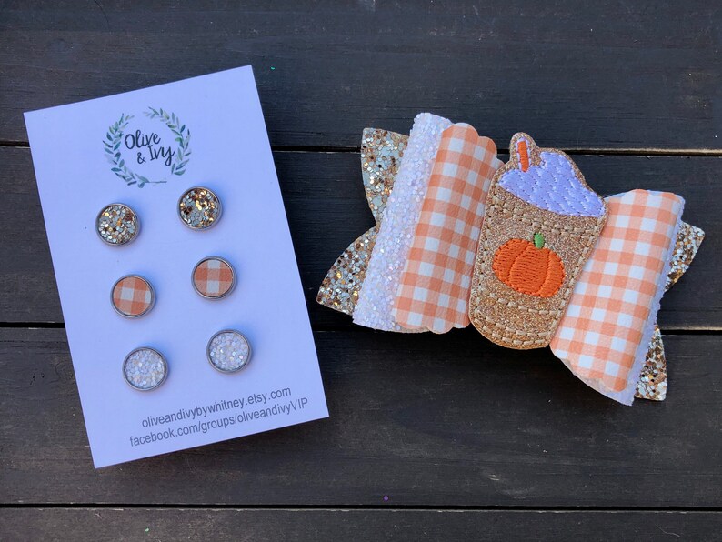Op de afbeelding: Set van drie 2,5 cm ronde haarstrikken met een pompoenspeceratte-ontwerp. De strikken zijn gemaakt van oranje en wit gingham-stof, gouden glitterstof en een paarse en oranje pompoenspeceratte-applicatie. De set bevat ook drie paar oorbellen met gouden glitter, witte glitter en oranje en wit gingham-stof.
