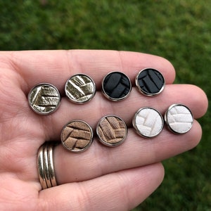 May include: Eight pairs of small, round, leather earrings in various colors, including black, white, brown, and gold. The earrings are displayed on a hand.