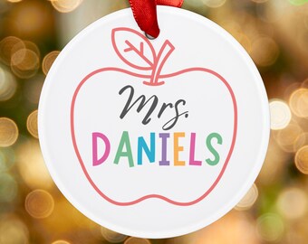 May include: White ceramic ornament shaped like a red apple with a green stem and leaf. The text "Mrs. Daniels" is written in colorful letters on the apple.