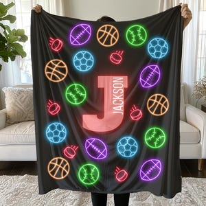May include: A black blanket featuring a neon design of various sports balls, including basketballs, soccer balls, baseballs, hockey pucks, and footballs. The letter "J" and the name "Jackson" are prominently displayed in the center.