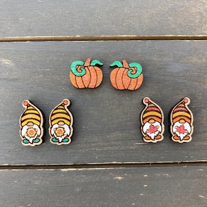 May include: A set of six wooden earrings featuring a fall theme. Two earrings are shaped like pumpkins with green stems and orange bodies. The other four earrings are shaped like gnomes with yellow and orange striped hats, brown beards, and red noses.