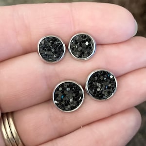 May include: Four black glitter stud earrings with silver settings. The earrings are displayed on a hand.