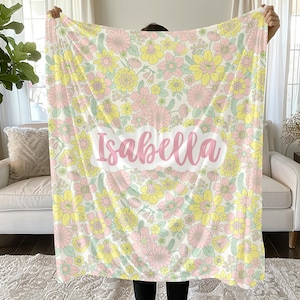 May include: A soft blanket featuring a repeating floral pattern in shades of pastel pink, yellow, and light green on a cream background. The name Isabella is printed in a large, pink, cursive script across a white cloud-shaped center panel.