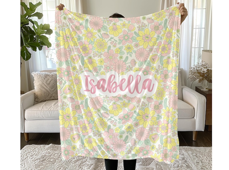 May include: A soft blanket featuring a repeating floral pattern in shades of pastel pink, yellow, and light green on a cream background. The name Isabella is printed in a large, pink, cursive script across a white cloud-shaped center panel.
