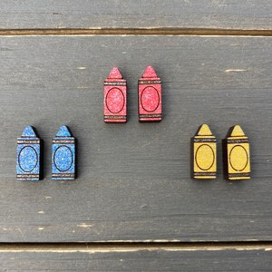 K&ouml;nnte beinhalten: Sechs Ohrringe in Form von Holzstiften in Rot, Blau und Gelb. Die Ohrringe sind alle gleich gro&szlig; und haben die gleiche Form und eine glitzernde Oberfl&auml;che.