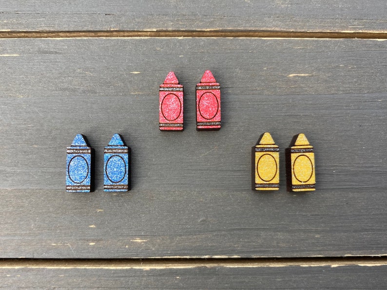 K&ouml;nnte beinhalten: Sechs Ohrringe in Form von Holzstiften in Rot, Blau und Gelb. Die Ohrringe sind alle gleich gro&szlig; und haben die gleiche Form und eine glitzernde Oberfl&auml;che.