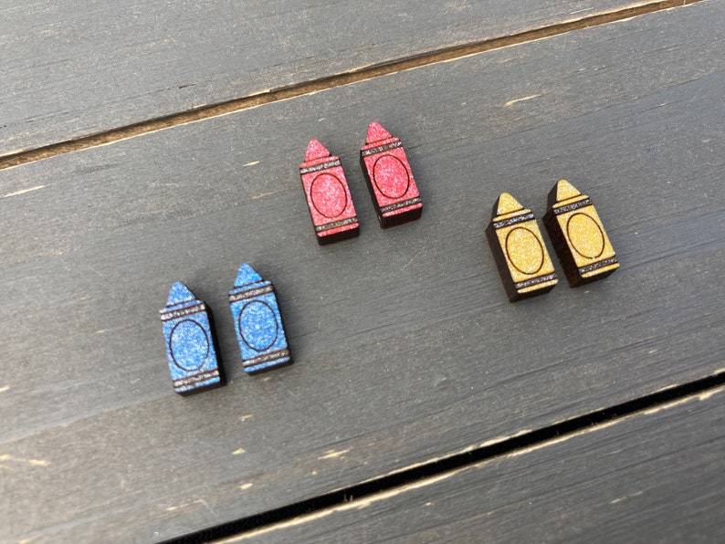 K&ouml;nnte beinhalten: Ein Set mit sechs Holz-Kreide-Ohrringen. Zwei rote, zwei blaue und zwei gelbe Kreiden mit Glitzer.