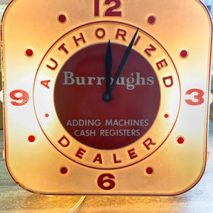 May include: A square, illuminated wall clock with a cream-colored border and a deep red center. The face features the text AUTHORIZED Burroughs DEALER, ADDING MACHINES, and CASH REGISTERS, with large red numerals at the 12, 3, 6, and 9 positions.
