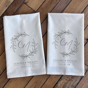 May include: Two off-white rectangular kitchen towels featuring a black printed design of a botanical wreath surrounding the letters C+M. Below the wreath, the text reads CLAUDIA & MICHAEL followed by the date 21.07.24 in a clean sans-serif font.