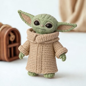 以下が含まれることがあります： 大きな耳と大きな目を持つキャラクターの手作りの編み人形。人形は緑色で、ベージュのローブを着ており、高さは約15cmです。SFシリーズで人気のキャラクターです。