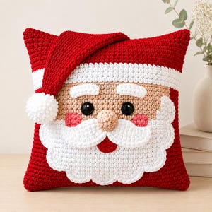 May include: A square crochet pillow featuring a Santa Claus face design. The pillow is primarily red with a white brim and a dangling red hat with a white pom-pom. It has a beige face, black button eyes, a beige nose, and a white beard.