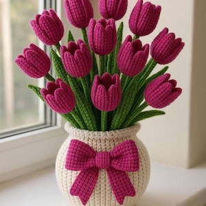 May include: A handmade crochet bouquet of magenta tulips in a cream-colored vase with a magenta bow. The tulips have green stems and leaves. The vase is sitting on a white windowsill.