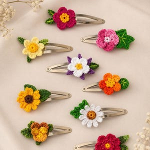 Könnte beinhalten: Eine Sammlung von neun silberfarbenen Metall-Haarspangen, jede mit einem einzigartigen gehäkelten Blumendesign in verschiedenen Farben wie Magenta, Gelb, Pink, Weiß, Lila, Orange und Braun, akzentuiert durch kleine grüne gehäkelte Blätter.