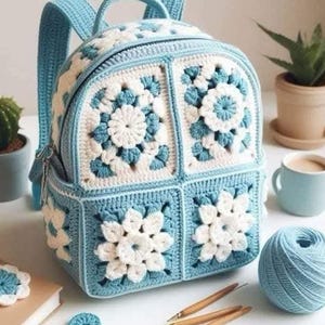 Op de afbeelding: Een handgemaakte gehaakte rugzak in lichtblauw en wit. De tas heeft een bloemen granny square ontwerp, een ritssluiting, een klein voorvak en verstelbare schouderbanden. De afbeelding toont ook haaknaalden en een bol garen.