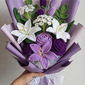 Puede incluir: Un ramo de crochet hecho a mano con lirios blancos, rosas moradas y ramitas tipo lavanda, envuelto en papel lila y atado con una cinta de encaje blanco. Un arreglo floral único y artesanal.