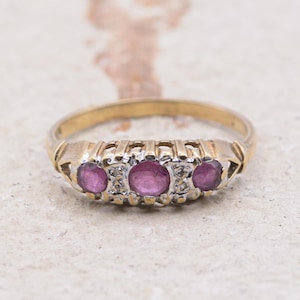 Puede incluir: Un anillo de oro con tres piedras preciosas rosas ovaladas dispuestas en fila. El anillo tiene una banda detallada con recortes decorativos y pequeñas piedras de acento. El anillo se muestra sobre una superficie neutra y texturizada.