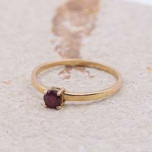 May include: A gold ring featuring a round, deep red gemstone. The ring has a slender band and a classic design. The gemstone is set in a four-prong setting.