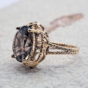 May include: A gold ring with a large, oval-shaped, smoky quartz gemstone set in a delicate, rope-like design.