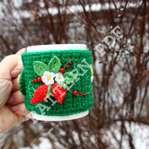 Puede incluir: Un calientavasos de ganchillo verde con un diseño de fresa y flores. El calientavasos está en una taza blanca.