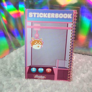 May include: A spiral-bound sticker book with a pink and purple arcade game design. The cover reads "STICKERBOOK" in white letters. A cartoon Shiba Inu hangs from a pink claw. The bottom has buttons that say "DOWN".