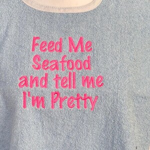 May include: Light blue terry cloth bib with a white trim around the neck. The bib features the embroidered phrase, "Feed Me Seafood and tell me I'm Pretty" in bright pink thread.