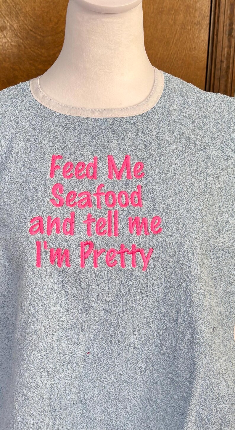 May include: Light blue terry cloth bib with a white trim around the neck. The bib features the embroidered phrase, "Feed Me Seafood and tell me I'm Pretty" in bright pink thread.