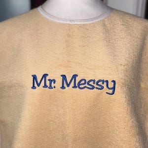 May include: A tan-colored sweatshirt with the embroidered text "Mr. Messy" in blue. The sweatshirt has a crew neck with a light-colored trim. The fabric appears soft and textured.