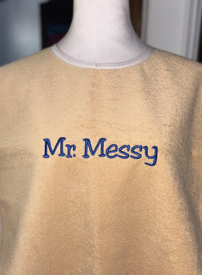 May include: A tan-colored sweatshirt with the embroidered text "Mr. Messy" in blue. The sweatshirt has a crew neck with a light-colored trim. The fabric appears soft and textured.
