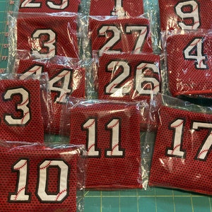 May include: A collection of red mesh fabric patches with white numbers and baseball stitching. Numbers include 23, 1, 9, 4, 3, 27, 26, 11, 17, and 10. Each patch is individually packaged in clear plastic.