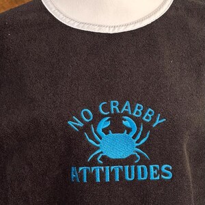 May include: Dark gray t-shirt with a white collar and the embroidered phrase "NO CRABBY ATTITUDES" in turquoise. The design includes a turquoise crab graphic.