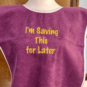 May include: A purple bib with white trim and the embroidered text "I'm Saving This for Later" in yellow. The bib is made of a soft, absorbent material, designed to catch spills and protect clothing.