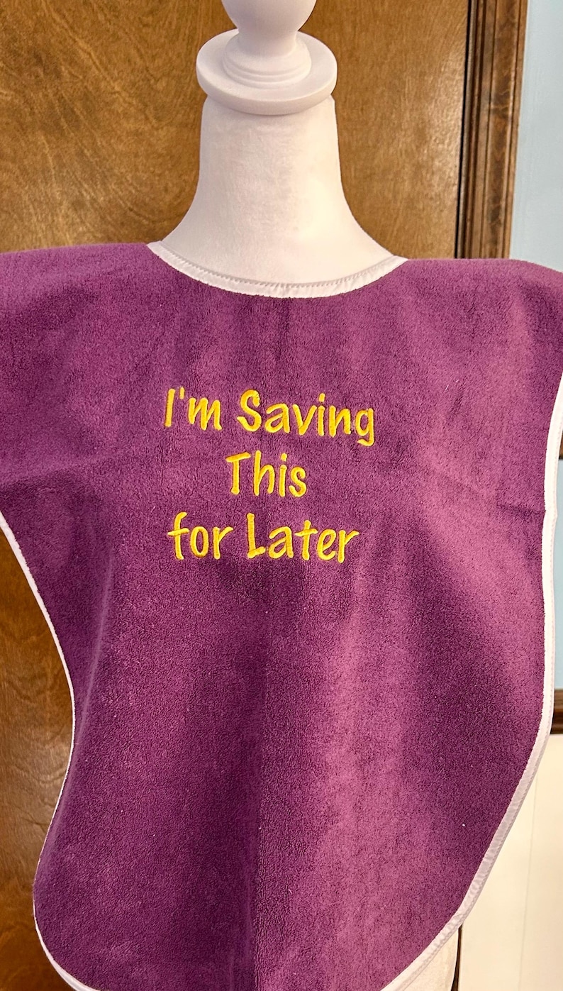 May include: A purple bib with white trim and the embroidered text "I'm Saving This for Later" in yellow. The bib is made of a soft, absorbent material, designed to catch spills and protect clothing.