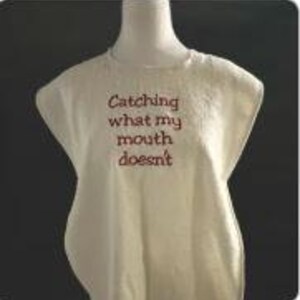 May include: Cream-colored sleeveless shirt with the embroidered text "Catching what my mouth doesn't" in a dark red color. The shirt is displayed on a mannequin against a dark background.