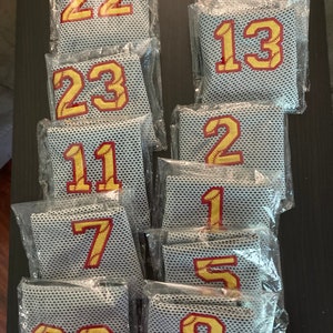 May include: A collection of mesh baseball jersey patches, each individually packaged. The patches feature large, gold-colored numbers with red baseball stitching, ranging from 00 to 23. The mesh material is a light gray color.