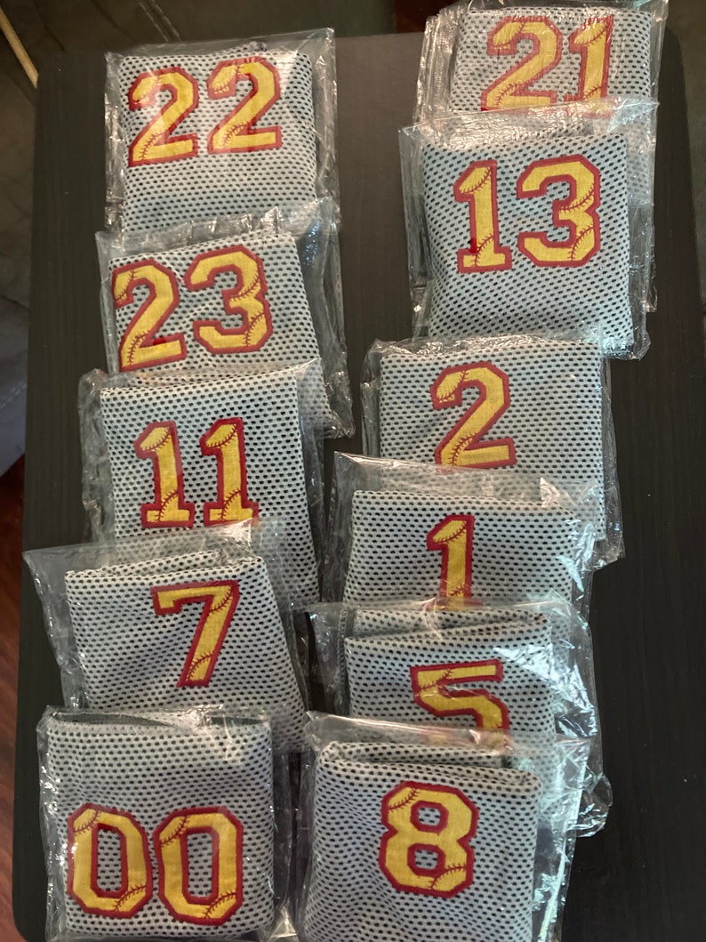 May include: A collection of mesh baseball jersey patches, each individually packaged. The patches feature large, gold-colored numbers with red baseball stitching, ranging from 00 to 23. The mesh material is a light gray color.