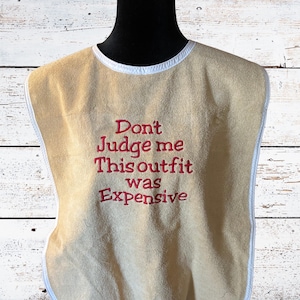 May include: Tan bib with white trim and the embroidered phrase "Don't Judge me This outfit was Expensive" in red. The bib is displayed on a black mannequin head against a light wood background.
