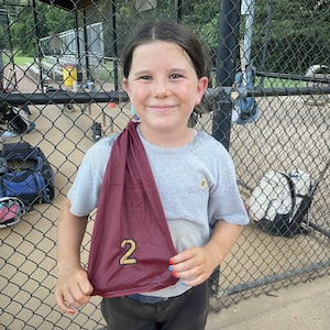 May include: A young person wearing a gray t-shirt and dark pants, holding a maroon mesh jersey with the number 2 in gold. The person is smiling in front of a chain-link fence, with a baseball field in the background.