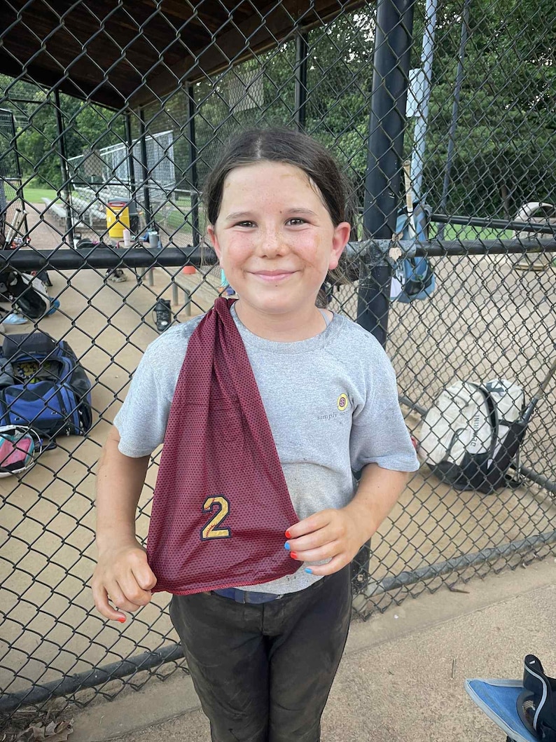 May include: A young person wearing a gray t-shirt and dark pants, holding a maroon mesh jersey with the number 2 in gold. The person is smiling in front of a chain-link fence, with a baseball field in the background.