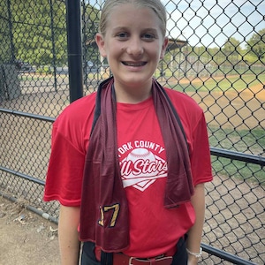 May include: A young person wearing a red t-shirt with the text "York County All Stars" and the number 17. They are wearing black pants and a red belt. A dark red towel is around their neck. The background shows a baseball field.
