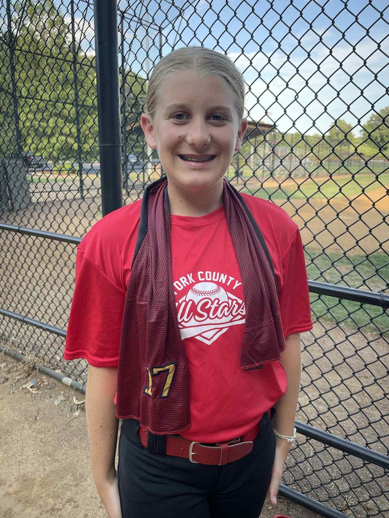 May include: A young person wearing a red t-shirt with the text "York County All Stars" and the number 17. They are wearing black pants and a red belt. A dark red towel is around their neck. The background shows a baseball field.
