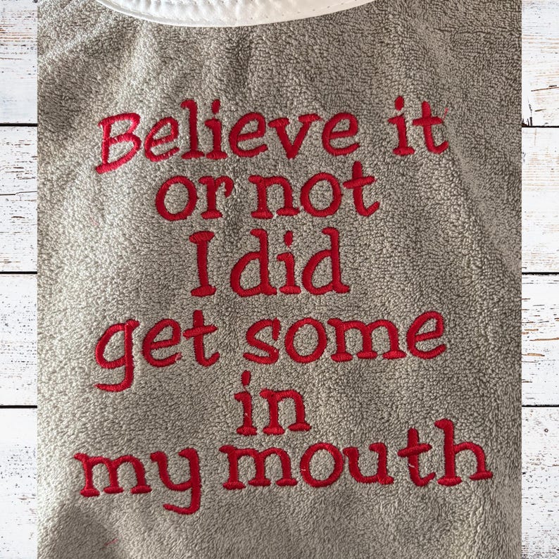 May include: A gray towel with the embroidered phrase "Believe it or not I did get some in my mouth" in red lettering. The towel has a white trim at the top and a textured surface.