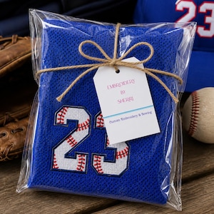 May include: A blue baseball jersey with the number 23 embroidered in white and red, packaged in clear cellophane and tied with twine. A tag reads "Embroidery by Sherri, Custom Embroidery & Sewing". A baseball and a blue baseball cap are in the background.