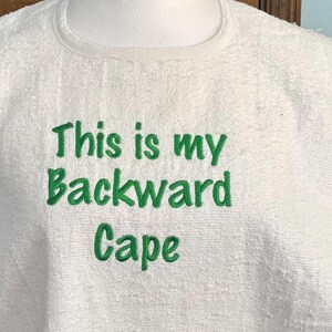 May include: White terry cloth cape with the embroidered text "This is my Backward Cape" in green. The cape is displayed on a white mannequin head and shoulders. The text is centered on the chest area.