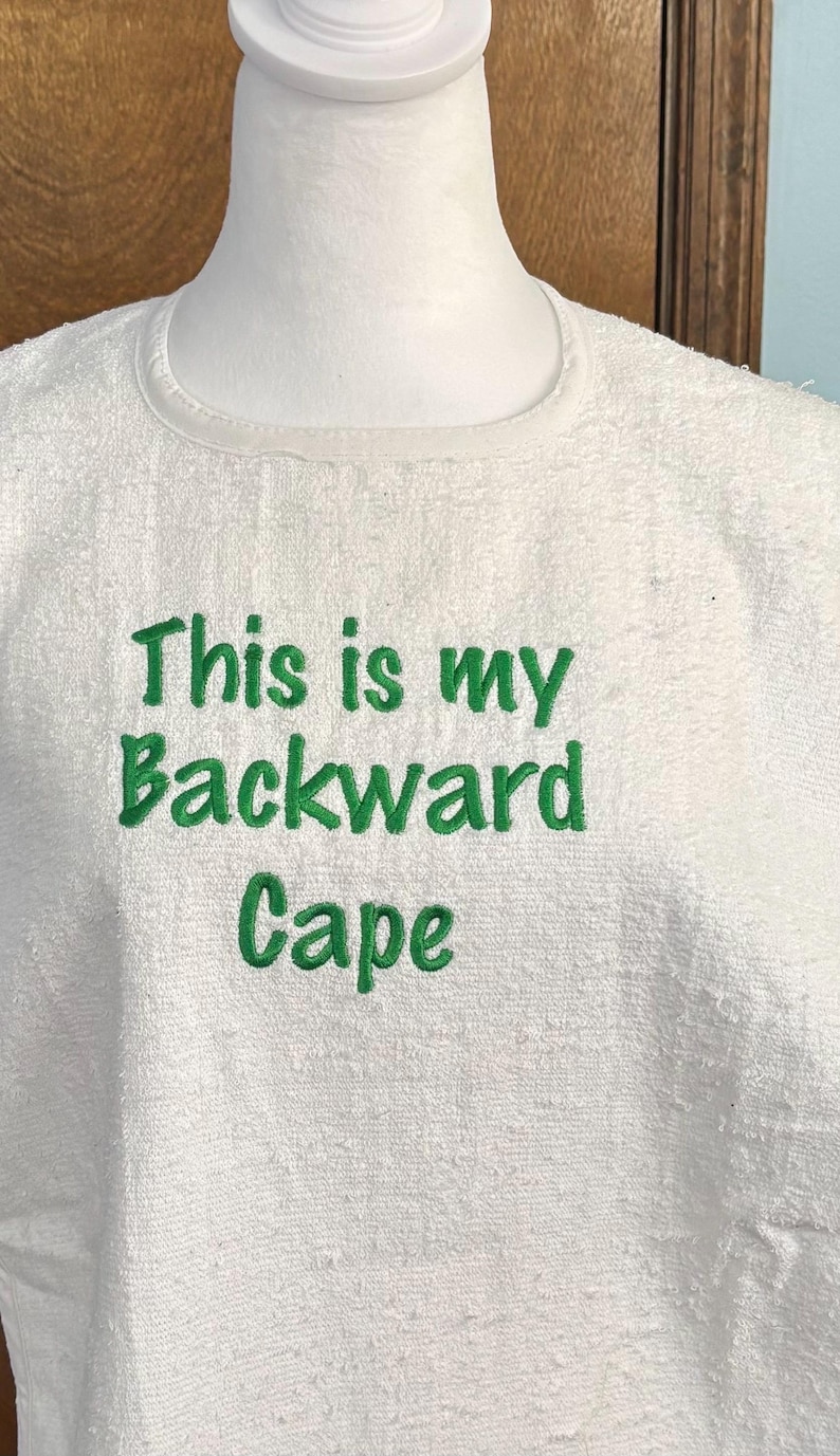 May include: White terry cloth cape with the embroidered text "This is my Backward Cape" in green. The cape is displayed on a white mannequin head and shoulders. The text is centered on the chest area.