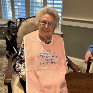 May include: A woman wearing a pink bib with the embroidered text "Don't Judge me This outfit was Expensive". The bib is around the woman's neck. She is sitting in a wooden chair.