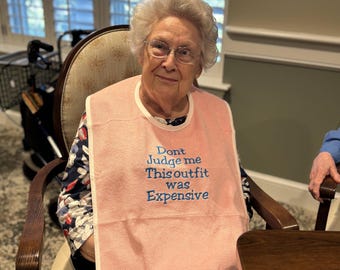 May include: A woman wearing a pink bib with the embroidered text "Don't Judge me This outfit was Expensive". The bib is around the woman's neck. She is sitting in a wooden chair.